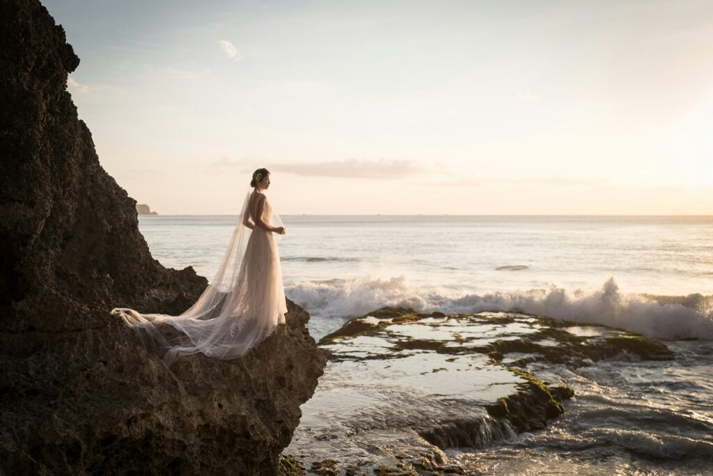 A bride standing on seaside rocks at sunset wearing a light wedding dress with a long veil