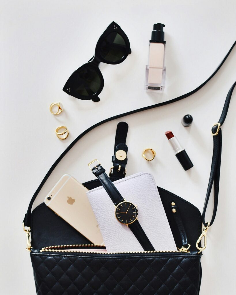 Flat lay of everyday fashion accessories including a handbag, sunglasses, makeup, phone, and watch for online shopping inspiration.