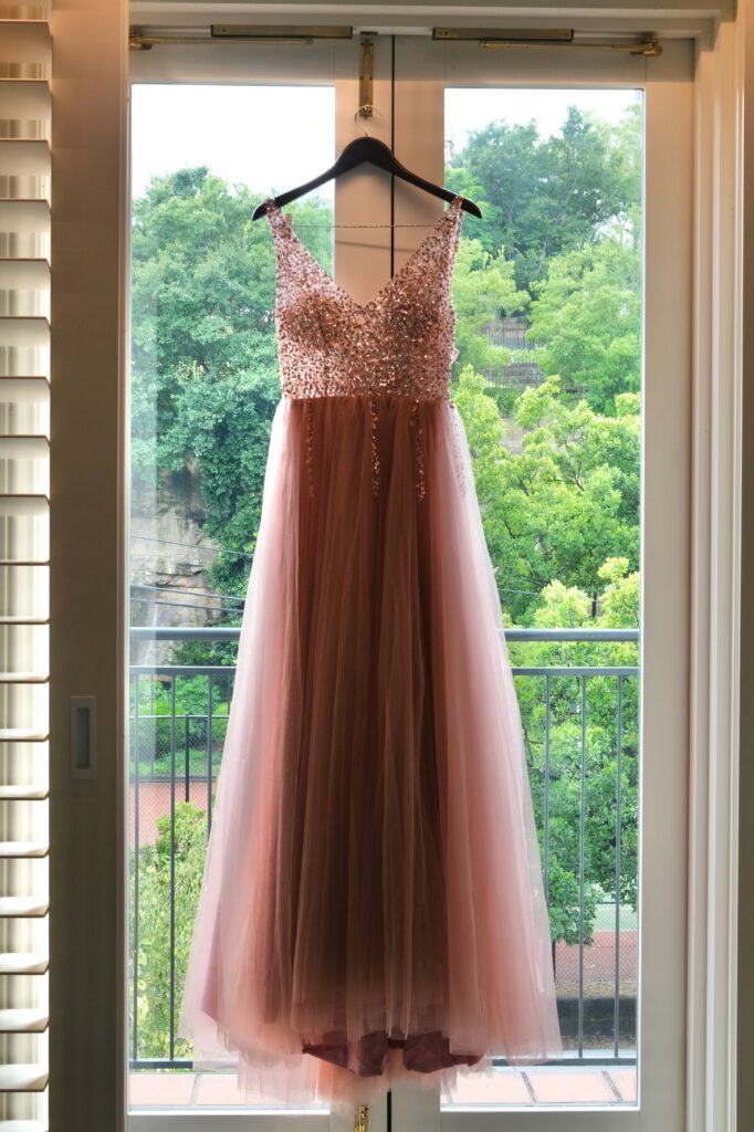 Blush pink prom dress with a sequined bodice and flowing tulle skirt hanging in front of a window