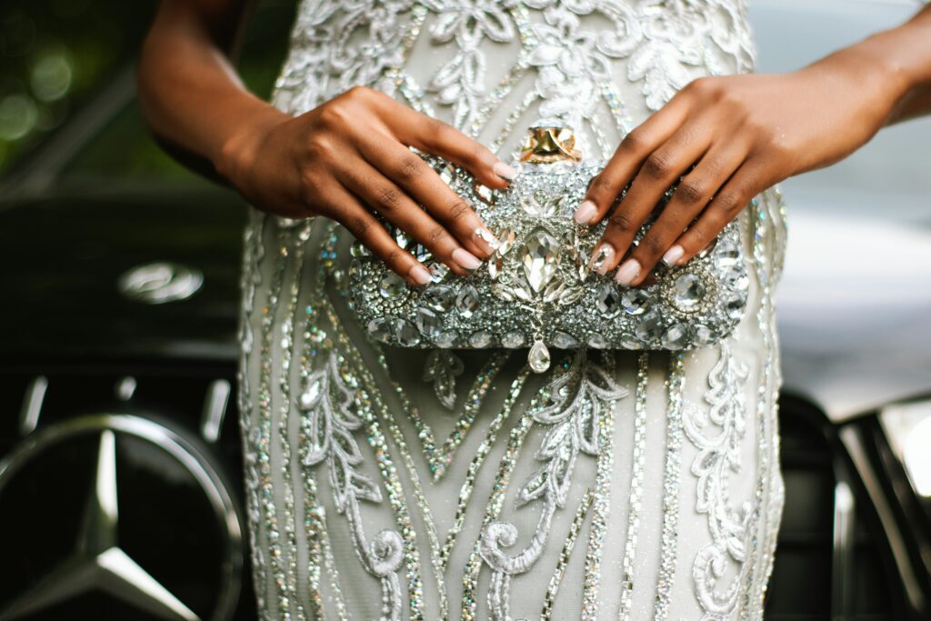Hands holding a silver crystal-embellished evening clutch against a white beaded formal gown